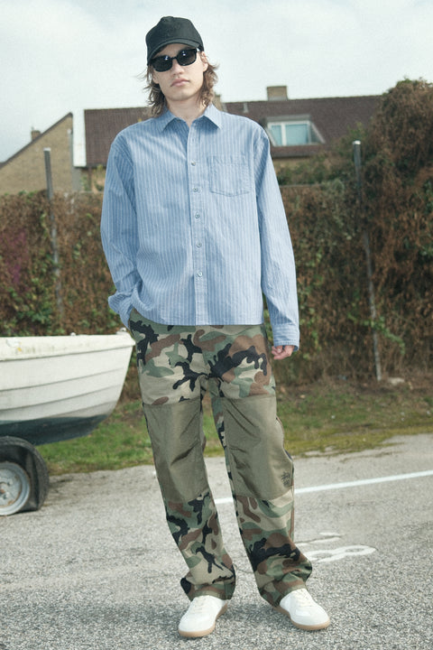 A person wearing a light blue shirt and camouflage pants is standing on a road with a boat and a house in the background.