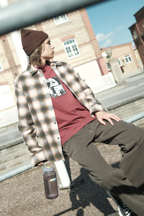 Person wearing a plaid shirt over a graphic t-shirt, sitting on a bench with a building in the background.